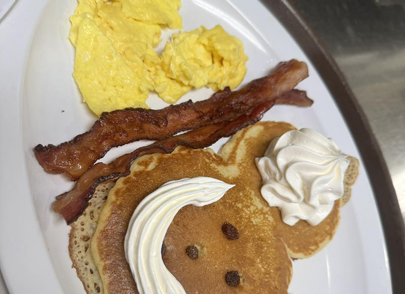 A plate of breakfast featuring two pancakes with whipped cream and a chocolate chip, scrambled eggs, and crispy bacon strips.