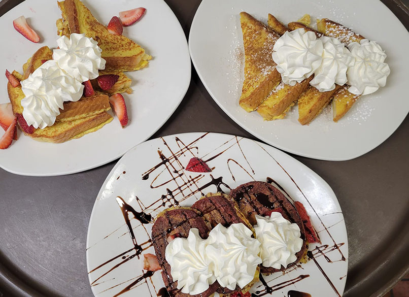 Three plates of French toast topped with whipped cream and fresh strawberries, drizzled with chocolate sauce on one plate.